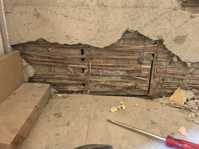 Skadad vägg där puts fallit av och exponerat underliggande träreglar och plankor, behov av reparation.
