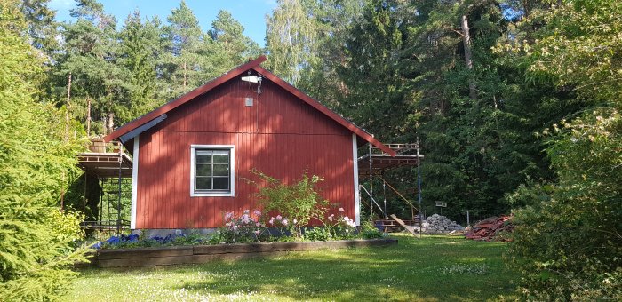 Rödmålat trähus med nymonterade vindskivor och byggställning intill, omgivet av gröna träd och en blomstrande trädgård.