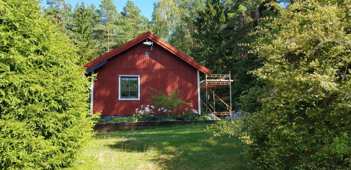 Rött hus med vita knutar och ett fönster, omgivet av grönska med en byggställning vid ena gaveln.