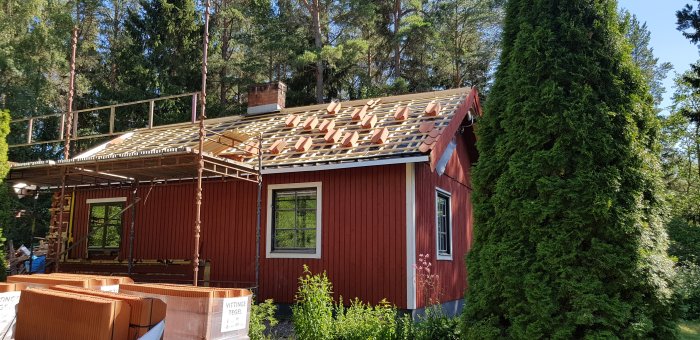 Röd stuga under takrenovering med uppmont. läkt och några takpannor provlagda, omgivet av grönska.