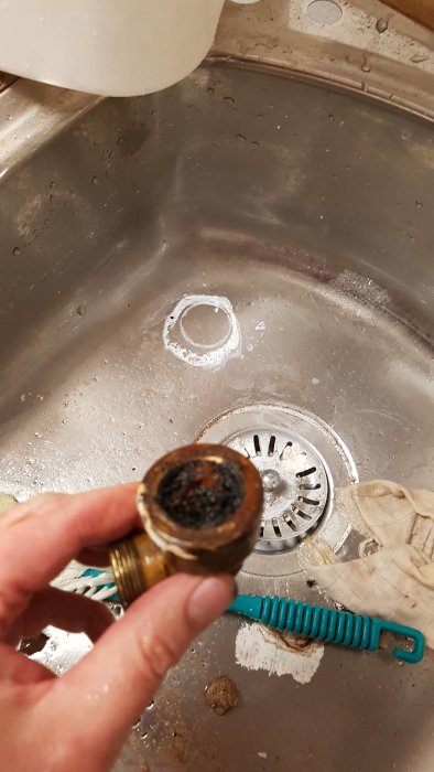 Hand håller en mässingskoppling med skadade gängor över en rostfri diskho med vattenstänk.