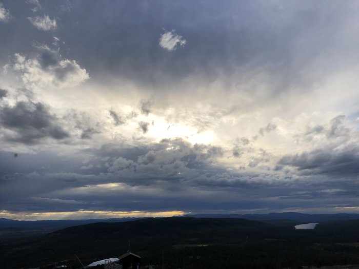 Bergsutsikt med dramatiska moln och solgenomsläpp över skogslandskap vid solnedgång.