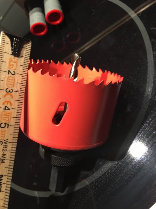 Röd hålsåg med borrhuvud som sticker ut cirka 15 mm, nära en linjal på en glansig yta.