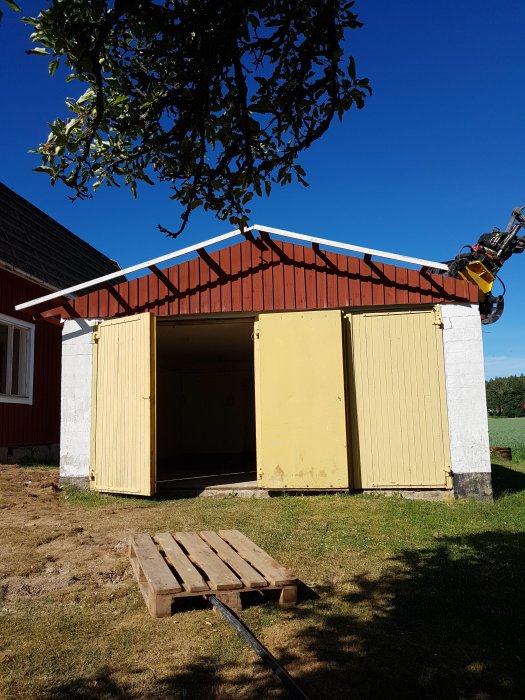 Ett gult garage med röd takfot och borttagna takstolar, öppna dörrar, under en klarblå himmel.