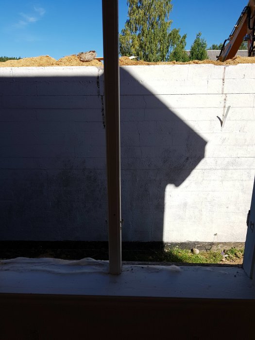 Utsikt från matsalsfönster med nära garagevägg och solskugga, tegel och rester på taket syns.