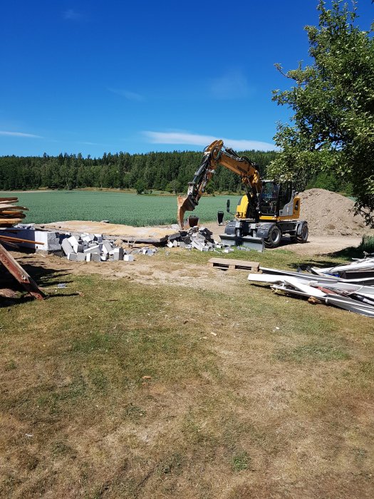 Grävmaskin river konstruktioner vid husrenovering med rivet material på marken och träd i bakgrunden.