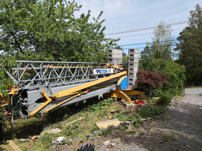 En gul och blå mobilkran med texten 'POTAIN' i en trädgård nära en byggarbetsplats.