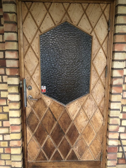 Ytterdörr i ek med rombisk mönster och en stor, svart, strukturerad glasruta, visande tecken på slitage och flagnad lack.
