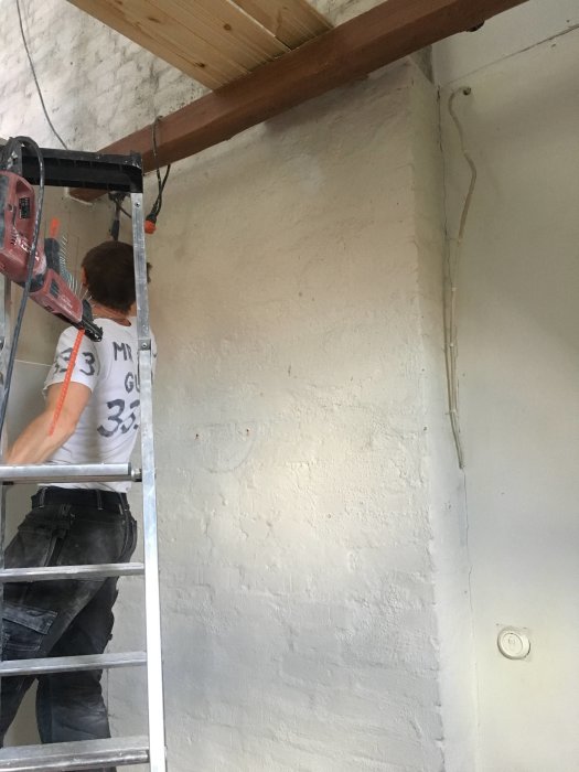 Person på en stege arbetar på en ojämn vitmålad tegelvägg med synliga missfärgningar och spår av reparationer.