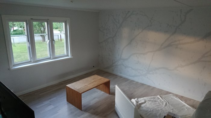 Vardagsrum med nylistade golv, möblerat med soffa och bord, och väggmålning som liknar trädgrenar.