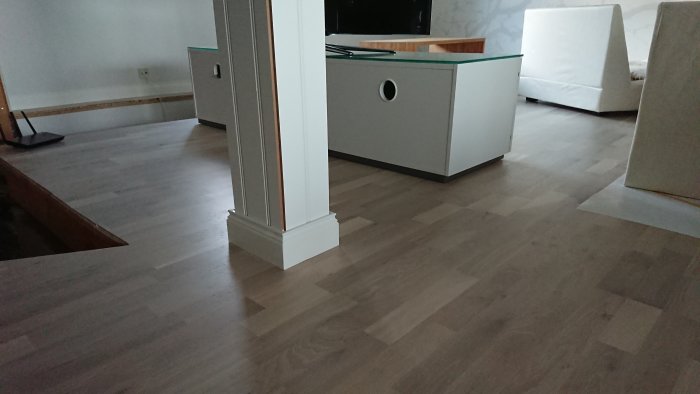 Nyinstallerade golvlistar i ett vardagsrum med laminatgolv och en vitmålad pelare, inredningsdetaljer i bakgrunden.