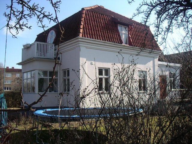 Vit villa med takkupa, röda takpannor och en balkong, framför en trampolin.