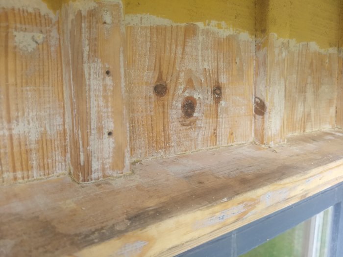 Träpanel med skrapade områden och kvarvarande tjocka färgrester vid skarvar och hörn, under gulmålad yta ovanför.