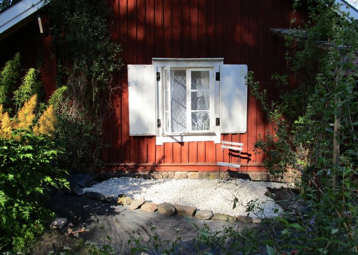 Nygrävd dräneringsränna med grus framför röd stuga och vit fönsterlucka omgiven av grönska.