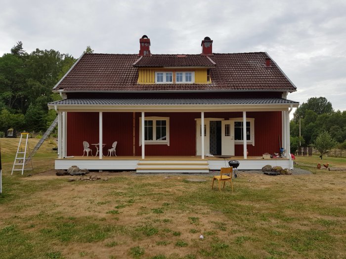 Ett rött hus med färdigt tak och oavslutad takavrinning, stege vid fasaden och uteplats med stolar.