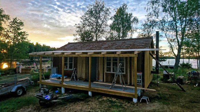 Hälften färdigbyggt verandatak på en trästomme i skymning med utsikt över en sjö och träd bakom.