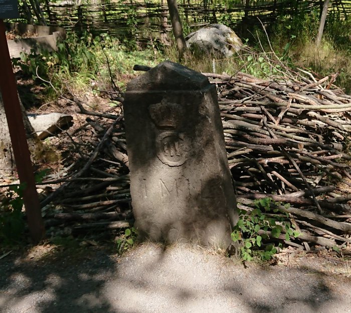 Gråsten med utskuret emblem och inskription framför en hög av vedträn i naturen.