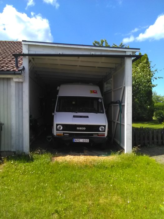 Husbil parkerad i en öppen carport bakom ett enfamiljshus, soligt väder.