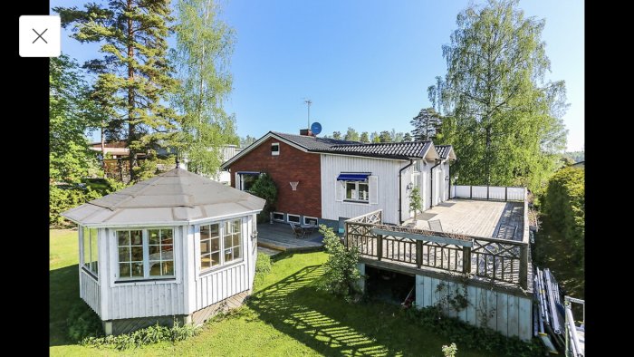 Ett rött tegelhus med ljusblå träpanelutbyggnad, terrass och grönskande trädgård i solljus.