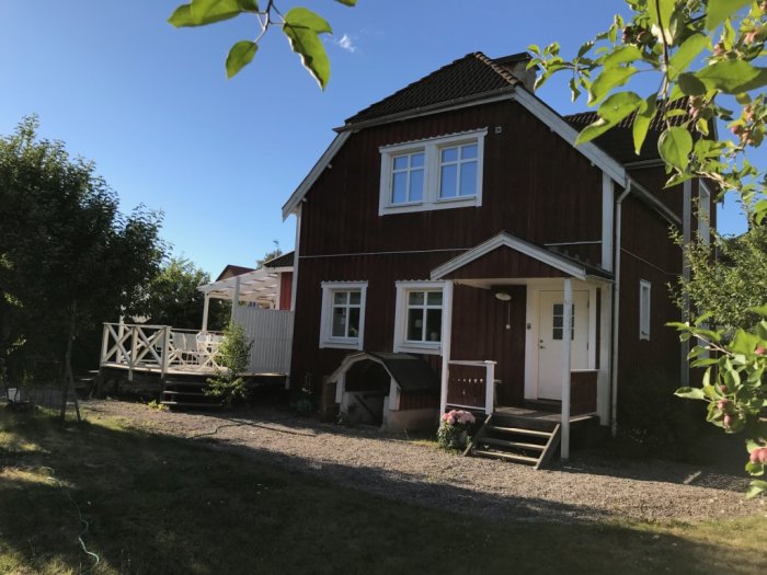 Rödmålat hus med farstukvist på plint och ett ordentligt tak, omgiven av grönska och en klarblå himmel.