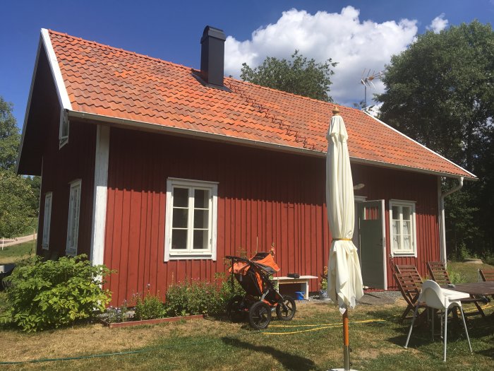 Röd trästuga med tegeltak och vita knutar under solig himmel, möjlig plats för tillbyggnad.