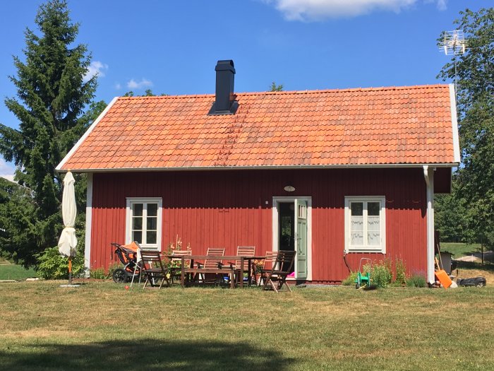 Röd trästuga med tegeltak, vita fönsterkarmar och en solig uteplats med bord och stolar.