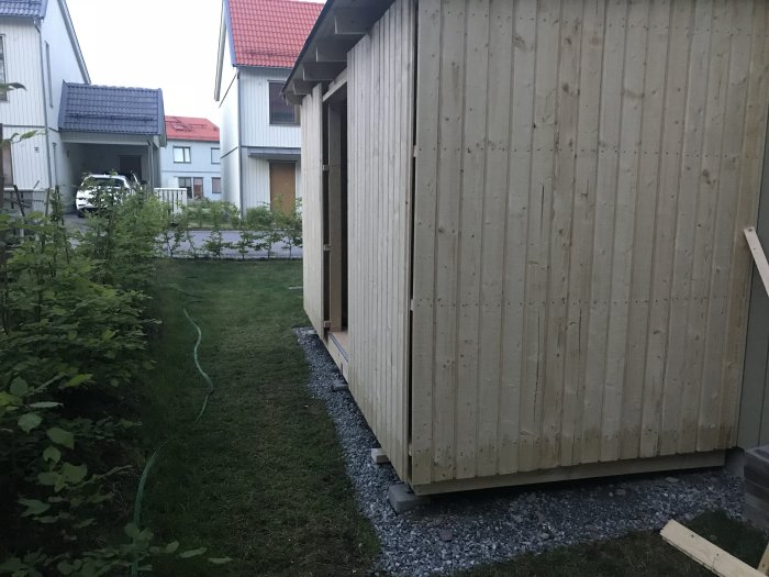 Obehandlad träpanel på ett nybyggt förråd vid skymning med grannhus i bakgrunden.