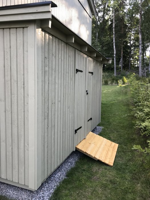 Nybyggd trärampe av spillvirke framför skjul med grundoljefärg, omgiven av gräs och grus.