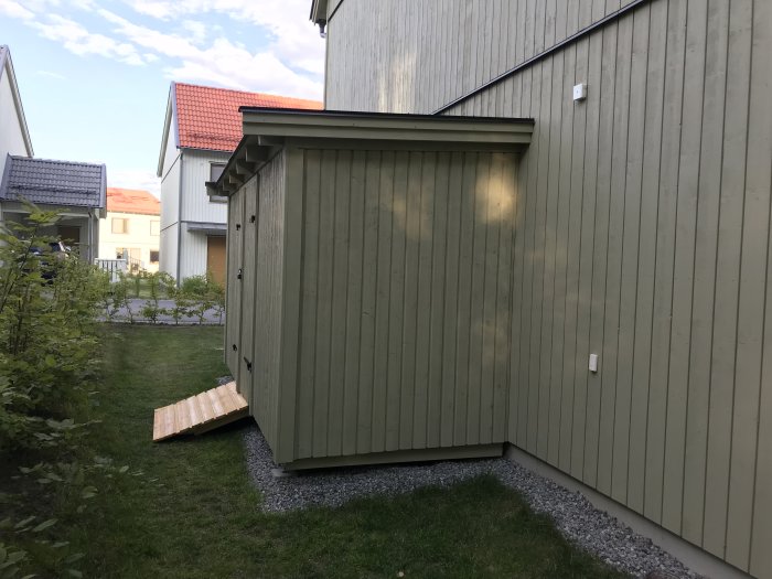 Nybyggd trärampramp av spillvirke framför en fristående förrådsbyggnad, omgiven av gräs och grusgång.