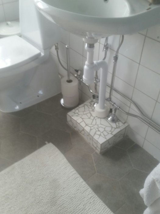 Badrum med vitt handfat och WC, och en handbyggd klack täckt av mosaik från spillplattor under handfatets rörinstallation.