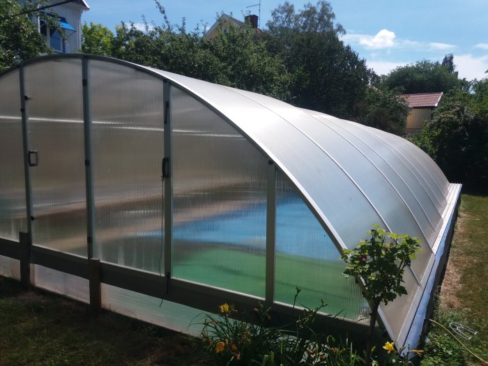 Halvrunt, höjt poolhus med transparent tak och väggar, omgivet av en trädgård och vegetation.