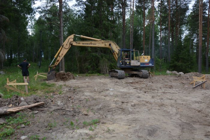 En grävmaskin och person på tomt med uppgrävd mark och synliga rännor för plintgjutning.