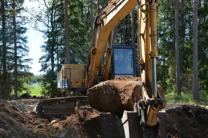 Grävmaskin som arbetar på en grävplats i skogen, stora jordhögar och uppgrävda rännor syns.