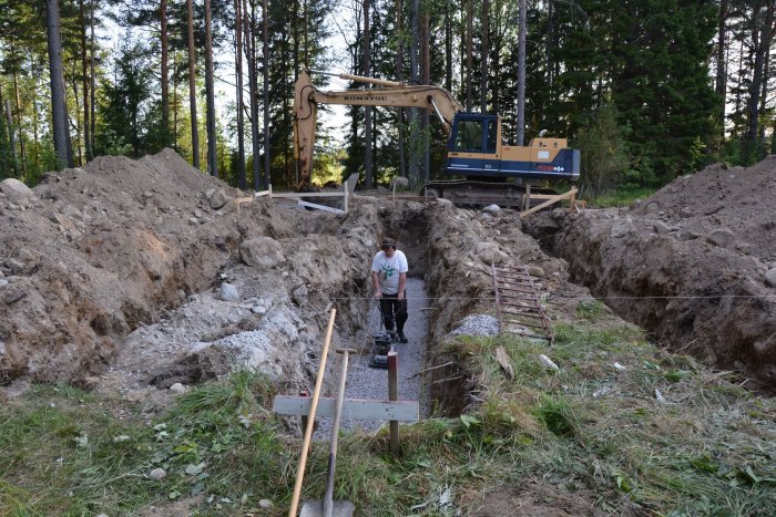 En person använder en jordstamp i en grävd ränna på en byggarbetsplats, med en grävmaskin och högar av uppgrävd jord i bakgrunden.