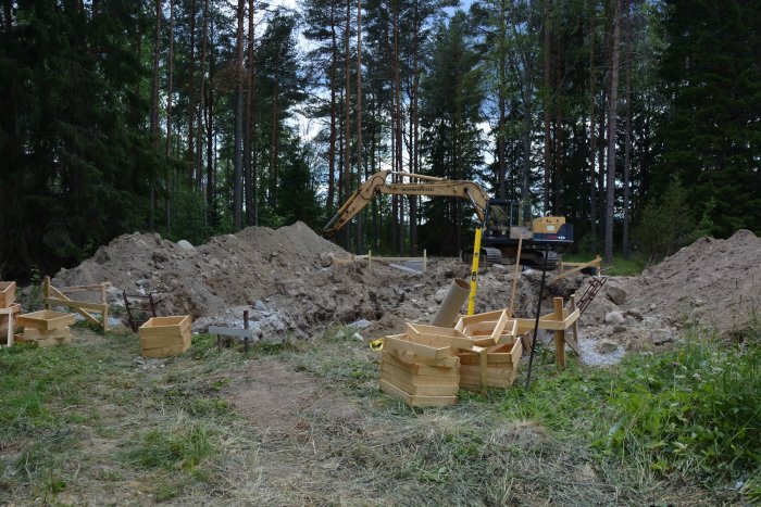 Byggarbetsplats med formar för gjutning av plintar, armeringsjärn och grävmaskin på ett skogstomt.