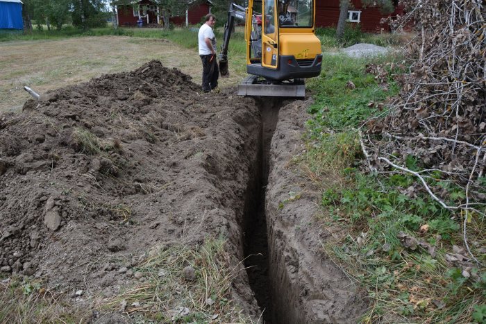 En grävmaskin och en person gräver en djup schakt för fundament med gräs och en röd byggnad i bakgrunden.