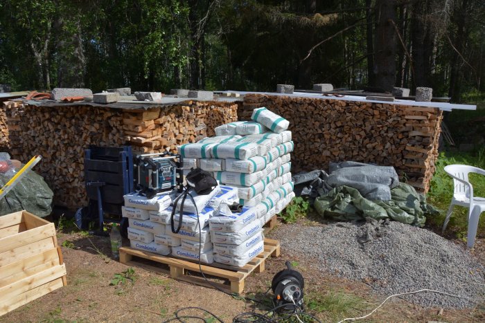 Byggplats med säckar av betong, armeringsjärn, vedstapel och byggmaterial.