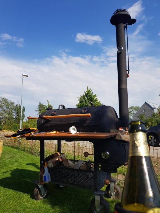 En utomhusgrill på hjul med vedhylla och grillverktyg på en solig dag, med blå himmel i bakgrunden och en champagneflaska i förgrunden.