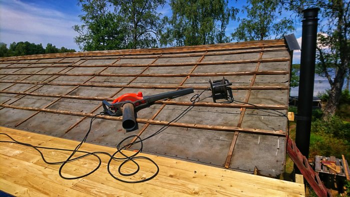 Tak under renovering med en lövsug och en lövblås liggandes på träreglar och betongpannor, med skog och sjö i bakgrunden.