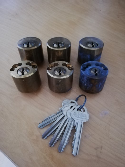 Tre par använda Assa 712 cylindrar och fem nycklar på ett träbord, i behov av putsning.