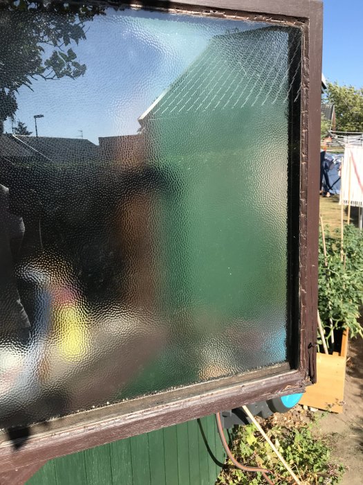 Slitet träfönster med frostat glas och skadad nedre kant, står utomhus med vegetation i bakgrunden.