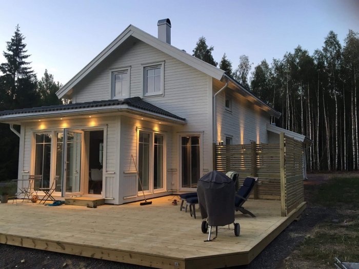Nybyggt trädäck med integrerad trall och staket vid vit tvåvåningshus i kvällsljus.
