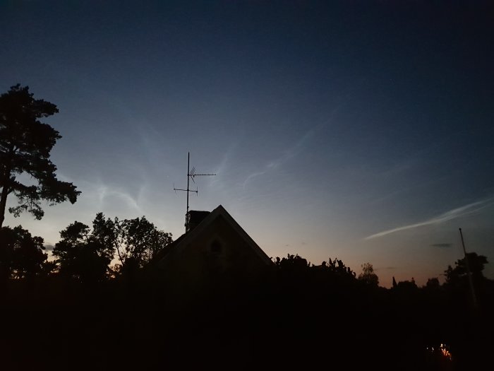 Nattbild över hus med antenntak och träd mot en mörkblå himmel med svagt ljusa molnformationer som påminner om norrsken.