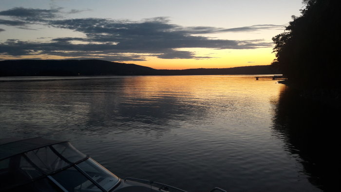 Midnattssol över lugnt vatten med silhuett av berg och båtens topp i förgrunden.