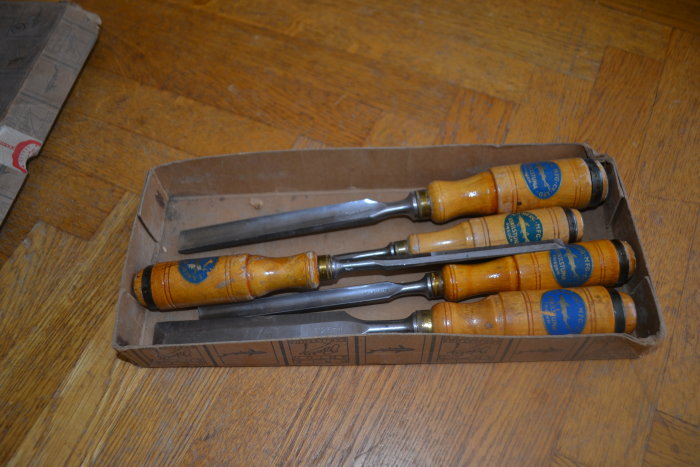 Fyra gamla E.A Berg-stämjärn i en brun papperslåda på trägolv, med trähandtag och blå etiketter.