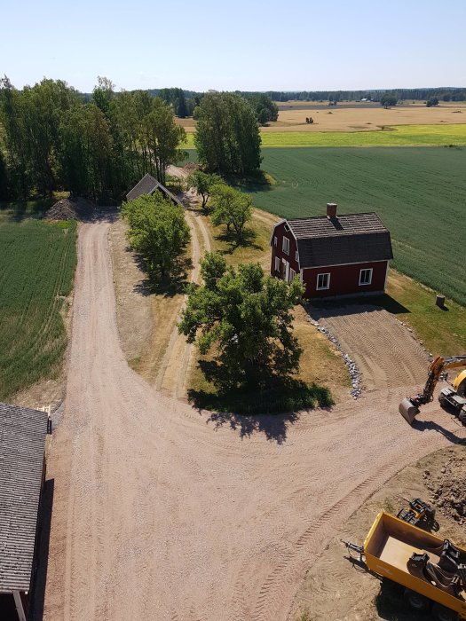 Vy mot söder över byggplats med rött hus, grusväg och omgivande fält.