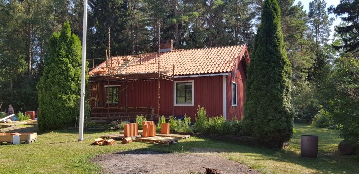 Rödfärgat hus med nylagda tegelpannor på taket, byggställning, material och nocktätning på marken.