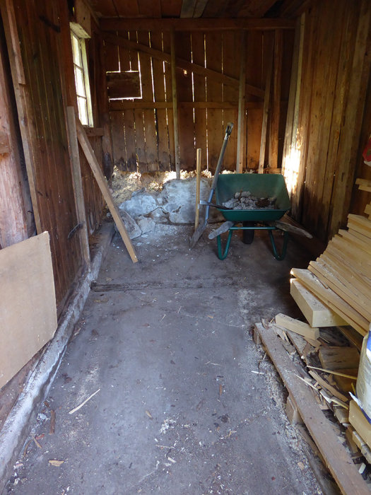 Vedbod efter rensning med synlig skadad grundmur, skottkärra och virkesstapel.