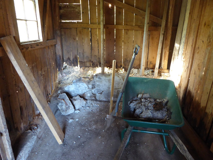 Inuti en vedbod med röjda rester och en delvis sönderslagen stenmur nära hörnet.