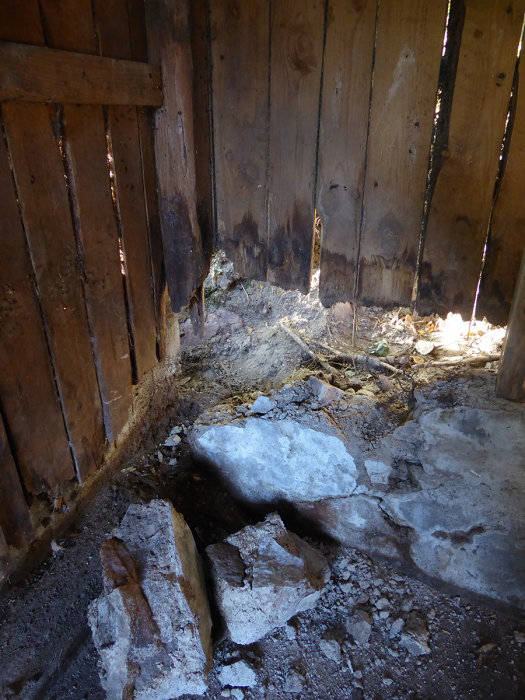 Sönderslagen grundmur med stora betongblock och spillror framför gammal trävägg i en vedbod, belyses av solljus.